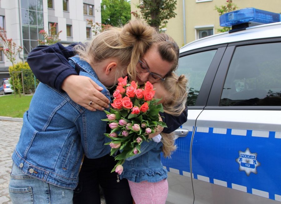 Mama - policjantka w mundurze trzyma w objęciach dwie dziewczynki, a w dłoni bukiet kwiatów. Obok zaparkowany jest policyjny radiowóz