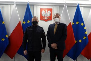 &quot;Umundurowany oficer Policji i mężczyzna w garniturze pozują do zdjęcia. Za nimi znajdują się flagi Polski i Unii Europejskiej oraz wisi godło Polski&quot;