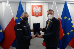 &quot;Umundurowany oficer Policji i stojący przy nim mężczyzna w garniturze trzymają pamiątkową tabliczkę. Obaj mężczyźni mają maseczki na twarzach. Za nimi znajdują się flagi Polski i Unii Europejskiej i wisi godło Polski&quot;