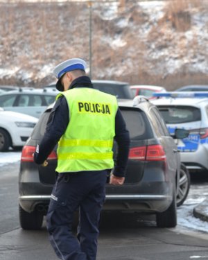 &quot;Policjant ruchu drogowego (widoczna biała czapka, kamizelka odblaskowa z napisem na plecach POLICJA) podchodzący do zatrzymanego do kontroli samochodu. W tle widać zimową aurę.&quot;