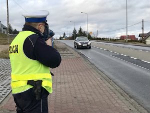 &quot;Policjant ruchu drogowego trzymając w ręku ręczny miernik prędkości mierzy prędkość jadącego po jezdni pojazdu&quot;