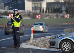 Policjant mierzy prędkość. z boku stoi radiowóz. W tle droga i przejeżdżające samochody.