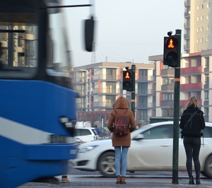 &quot;W centralnej części kadru widać dwie osoby piesze czekające przed przejściem dla pieszych na zmianę sygnału czerwonego zakazującego wejścia. W tle przejeżdżające po jezdni pojazdy. Z lewej strony kadru widać przejeżdżający tramwaj. Pogoda słoneczna, ubiór pieszych wskazuje na czas zimowy.&quot;