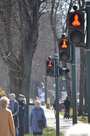 &quot;przez przejście dla pieszych z sygnalizacją świetlną nadawany kolor czerwony, przechodzą piesi.  Po drugiej stronie jezdni na chodniku stoi policjantka ruchu drogowego&quot;