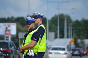 &quot;Dwóch policjantów ruchu drogowego stoi przy drodze nadzoruje ruch. W tle ruch uliczny słoneczna letnie pogoda&quot;