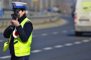 &quot;Policjantka ruchu drogowego mierzy prędkość przy drodze ręcznym miernikiem. W tle perspektywa drogi i ruch samochodów&quot;