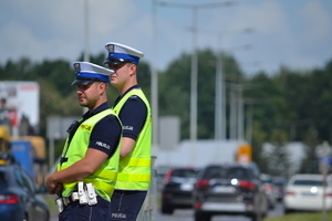 &quot;Dwóch policjantów ruchu drogowego stoi przy drodze i nadzoruje ruch. W tle widać ruch uliczny&quot;