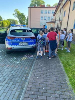 Dzień Dziecka w Nowym Brzesku. Na zdjęciach widać zawieszone na ogrodzeniu plakaty przedstawiające dzień dziecka. Widać również radiowóz a wokół niego zgromadzone dzieci i dzielnicowego, który opowiada.