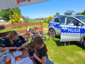 Dzień Dziecka Ostrów. Na zdjęciach widać, w którym siedzą dzieci oraz siedzącą przy stole policjantkę robiącą odciski palców. jedna z dziewczynek rozwiązuje rebus.