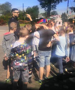 Dzień Dziecka w Nowym Brzesku. Na zdjęciach widać zawieszone na ogrodzeniu plakaty przedstawiające dzień dziecka. Widać również radiowóz a wokół niego zgromadzone dzieci i dzielnicowego, który opowiada.